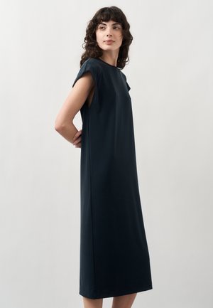 Γυναίκα με σγουρά μαλλιά που φοράει ένα αμάνικο σκούρο μπλε μεσαίο φόρεμα, στέκεται με τα χέρια στην πλάτη, κοιτώντας ελαφρώς στο πλάι.