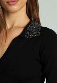 Schwarzer Pullover mit V-Ausschnitt und einem Kragen, der mit schwarzen Perlen verziert ist, wobei ein strukturiertes Designdetail im Schulterbereich hervorgehoben wird.