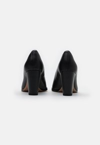 Escarpins en cuir noir à talons hauts avec bout pointu, large talon carré et texture lisse, présentant un design minimaliste sans détails supplémentaires.