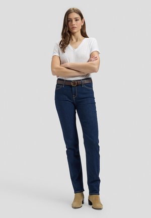 Ung kvinde med langt brunt hår iført en hvid T-shirt med V-hals, mørkeblå jeans med brunt bælte og lysebrune støvler med hæl, stående med armene over kors.