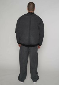 Zwarte, oversized pufferjack met een geribde zoom, gedragen met bijpassende donkergrijze broek met wijde pijpen, vanaf de achterkant gezien.