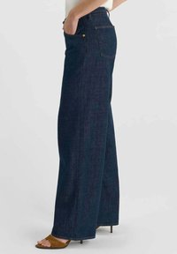 Wijd uitlopende donkerblauwe spijkerbroek gemaakt van denim. Voorzien van voorzakken, een hoge taille en zichtbare stiksel details. Geaccentueerd met gouden hardware.