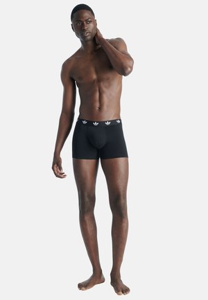 Młody mężczyzna stojący boso, ubrany w czarne bokserki Adidas, ręką dotykający szyi, lekko patrzący na bok na białym tle.