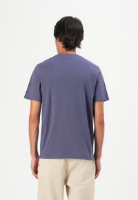 Hombre de pie de espaldas, vistiendo una camiseta azul de manga corta y pantalones beiges, contra un fondo blanco.