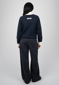 Femme aux cheveux noirs portant un pull bleu marine avec des poignets blancs et un col haut, vêtue d’un pantalon large bleu marine à rayures, vue de dos.
