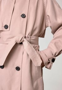 Ljusrosa trenchcoat med dubbla svarta knappar, bälte i midjan och manschettdetaljer med söm.
