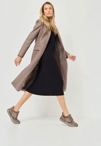 Manteau long marron sur une robe noire côtelée avec un ourlet au niveau des genoux. Baskets marron et en maille avec lacets. Fond clair, mannequin en mouvement.