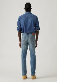 Cămașă din denim de culoare albastru deschis, cu un detaliu pe piesa din spate, asortată cu blugi albaștri decolorati, având o etichetă din piele la talie și picioare drepte.