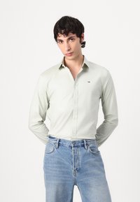 Cămașă butonată de culoare verde deschis, cu guler ascuțit, mâneci lungi și un accent cu un mic logo. Puritată cu blugi din denim albastru.