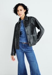 Svart skinnjakke med en glatt tekstur, knappefront og klassisk krage; lagd over en denimskjorte og flagrende jeans.