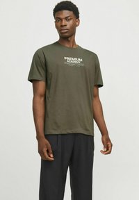 Olivno zelena bombažna majica s krojem ovratnika, kratkimi rokavi in belim tiskanim napisom "PREMIUM ACADEMY" na prsih. Črne hlače so oblečene spodaj.