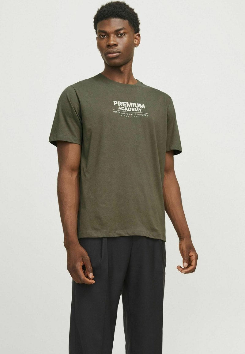Olivno zelena bombažna majica s krojem ovratnika, kratkimi rokavi in belim tiskanim napisom "PREMIUM ACADEMY" na prsih. Črne hlače so oblečene spodaj.