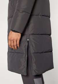 Manteau gris matelassé avec un fini mat, des coutures horizontales et une fermeture éclair sur le côté. Le tissu est lisse et légèrement réfléchissant.