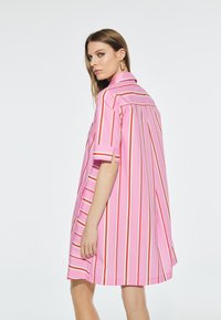 Růžové šaty s krátkým rukávem a vertikálními bílými a červenými pruhy. Mají límeček, volný střih a mírně zakřivený lem.