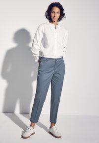 Camicia bianca a maniche lunghe con colletto arricciato, abbinata a pantaloni su misura azzurri e scarpe da ginnastica bianche con suola marrone.