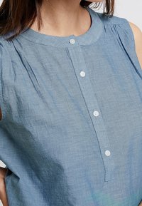 Blouse bleue sans manches avec un col rond. Présente des plis verticaux et une rangée de boutons sur le devant. Fabriquée en tissu léger.