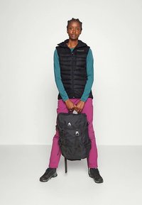 Crna prsluk s naletima, duge tirkizne rukave, ružičaste hlače, drži crni ruksak s logotipom planine, s podesivim trakama i patentnim zatvaračima.