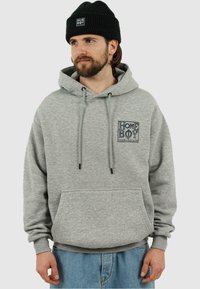 Felpa grigia con tasca frontale, coulisse e logo "HOME BOY". Abbinata a pantaloni di jeans azzurri chiari e a un berretto nero a coste.