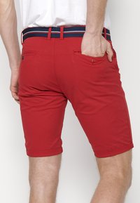 Shorts rouges avec un ourlet replié, en tissu léger. Présente une ceinture rayée bleu marine et blanche. Deux poches arrières sont visibles.