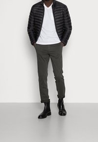 Giacca a vento nera da uomo, T-shirt bianca a V, pantaloni verde oliva e stivali neri alla caviglia. Texture liscia con design imbottito e polsini arrotolati.