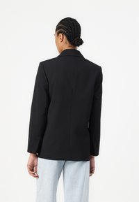 Blazer nero su misura con rever a punta, maniche lunghe e una texture liscia, abbinato a pantaloni a gamba larga azzurri.