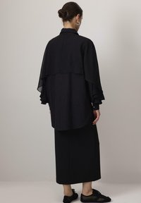 Black layered blouse with a lightweight, sheer overlay and ruffled sleeve accents, matched with a long black skirt and black footwear.
