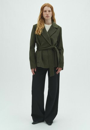 Femme aux longs cheveux roux portant un manteau ceinturé vert foncé, un haut blanc et un pantalon large noir, debout devant un fond blanc uni.