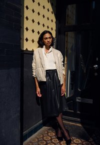 Femme portant une doudoune beige, un chemisier blanc, une jupe midi texturée noire, des collants noirs et des talons noirs, appuyée contre un mur et une porte à motifs.