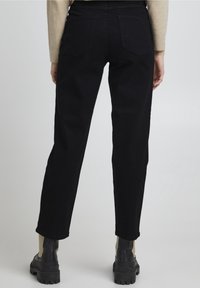 Pantalons en denim noir avec une coupe droite, design à cinq poches, couture visible et taille mi-haute. Portés avec des bottes noires épaisses.