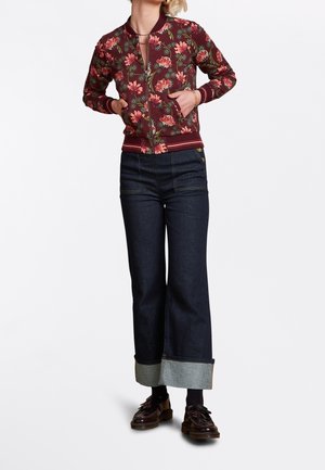 Woman wearing a red floral bomber jacket with hands in pockets, paired with dark wide-leg jeans with large cuffs and brown loafers.