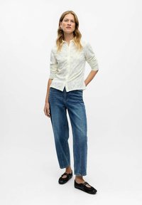 Donna in piedi su uno sfondo bianco, con una blusa bianca a maniche lunghe in pizzo, jeans blu e scarpe con cinghietti nere.