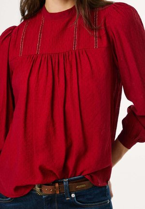 Vrouw draagt een gestructureerde rode blouse met pofmouwen en decoratieve verticale kanten strepen, gecombineerd met blauwe spijkerbroek en een bruine riem.