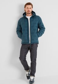 Teal puffer jacket with a hood, quilted texture, and zip closure. Worn over a white t-shirt, paired with dark jeans and blue sneakers.