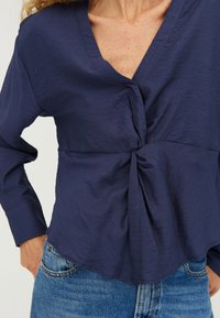 Maglietta blu navy con scollo a V e maniche lunghe, caratterizzata da un dettaglio anteriore annodato e un tessuto leggermente testurizzato. Abbinata a jeans in denim blu.