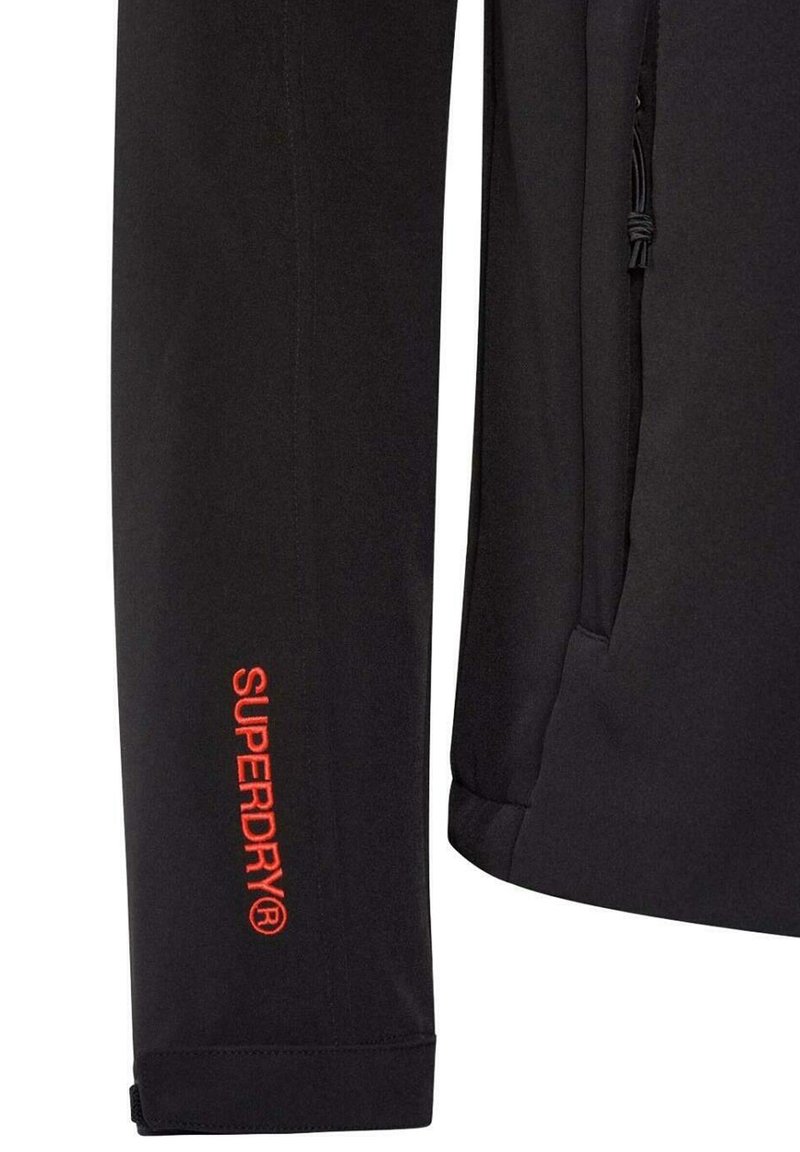 Zwarte buitenjack met een slank ontwerp, voorzien van een rode "SUPERDRY" logo op de mouw en elastische manchetten voor een getailleerde uitstraling.