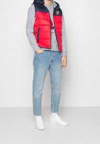 Gilet imbottito rosso e navy con zip davanti, indossato sopra un felpa grigia. Jeani azzurri e sneaker bianche completano l'outfit.