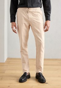 Helle beige Baumwollhose mit elastischem Kordelzugbund, schmaler Schnitt und Seitentaschen, kombiniert mit schwarzen Slippern.