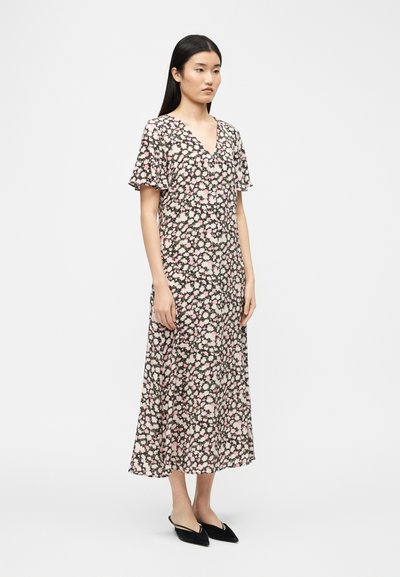 Γυναίκα που στέκεται φορώντας μαύρο μεσαίο φόρεμα με floral μοτίβο σε ροζ και λευκά λουλούδια, με λαιμόκοψη σε σχήμα V, κοντά μανίκια με βολάν και μαύρα slingback παπούτσια με μυτερή μύτη.