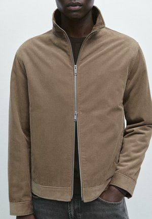 Un homme portant une veste en velours côtelé brun clair avec une double fermeture éclair sur une chemise sombre, avec une main dans la poche de son jean.