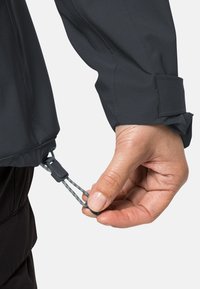 Zwarte jas met een gestructureerde afwerking, met een trekkoordmechanisme aan de zoom, vastgehouden in een hand voor aanpassing, met een manchet aan de mouw.