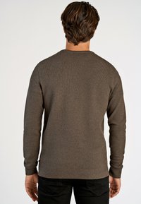 Ruskea neulepusero, jossa on teksturoitu neule, pyöreä pääntie ja pitkät hihat, joissa on ribbireunat. Yhdistetty tummaan housuiseen.