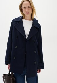 Palton trench din tweed bleumarin, cu rever mari și patru butoane frontale. Modelul ține o geantă maro, purtând un top alb și pantaloni din denim.