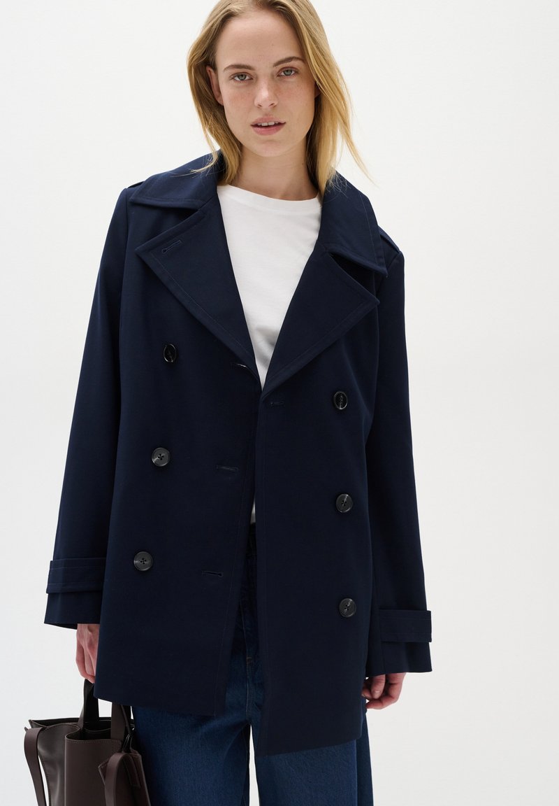 Palton trench din tweed bleumarin, cu rever mari și patru butoane frontale. Modelul ține o geantă maro, purtând un top alb și pantaloni din denim.