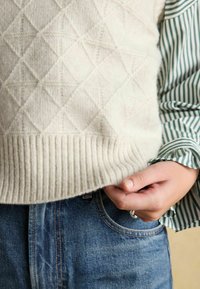 Cremefarbener, diamantgestickter Strickpullover mit geripptem Saum, getragen über einem grün-weiß gestreiften Hemd, kombiniert mit blauen Jeans.