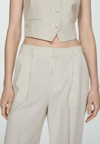 Chaleco corto de rayas beige con dos botones, combinado con pantalones de tiro alto con pliegues. Ambos prendas cuentan con una tela ligera y texturada.