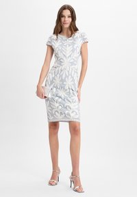 Hellblaues Kleid mit weißer Blumenstickerei, taillierte Form, kurze Ärmel und gewellter Saum. Model hält eine silberne Clutch und trägt silberne High Heels.