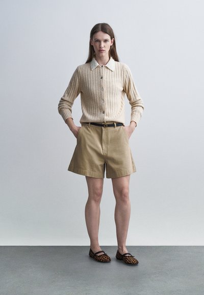Femme debout, mains dans les poches, portant un pull côtelé beige avec col blanc, un short kaki, une ceinture noire et des ballerines à imprimé léopard.