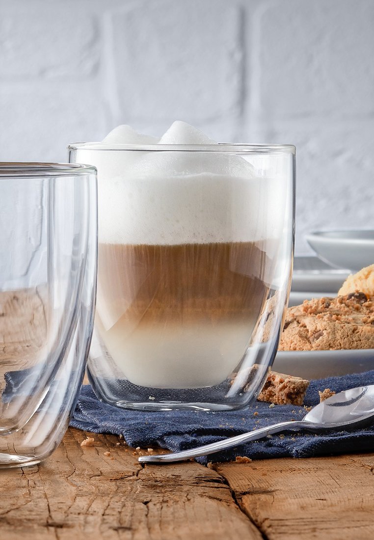 Taza de café de vidrio de doble pared que presenta capas de café marrón y espuma blanca. Acompañada de una cuchara de plata, una galleta y un paño azul.
