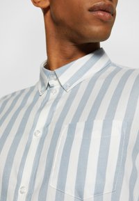 Camicia a righe blu chiaro e bianca con colletto button-down, taschino singolo e tessuto di cotone liscio. Presenta bottoni lucidi.