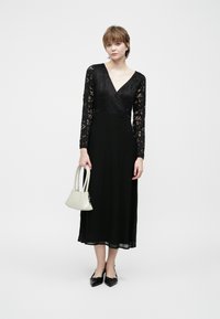 Svart blondekjole med lange ermer, V-hals og flytende skjørt. Kombinert med en liten hvit håndveske og svarte spisse sko.